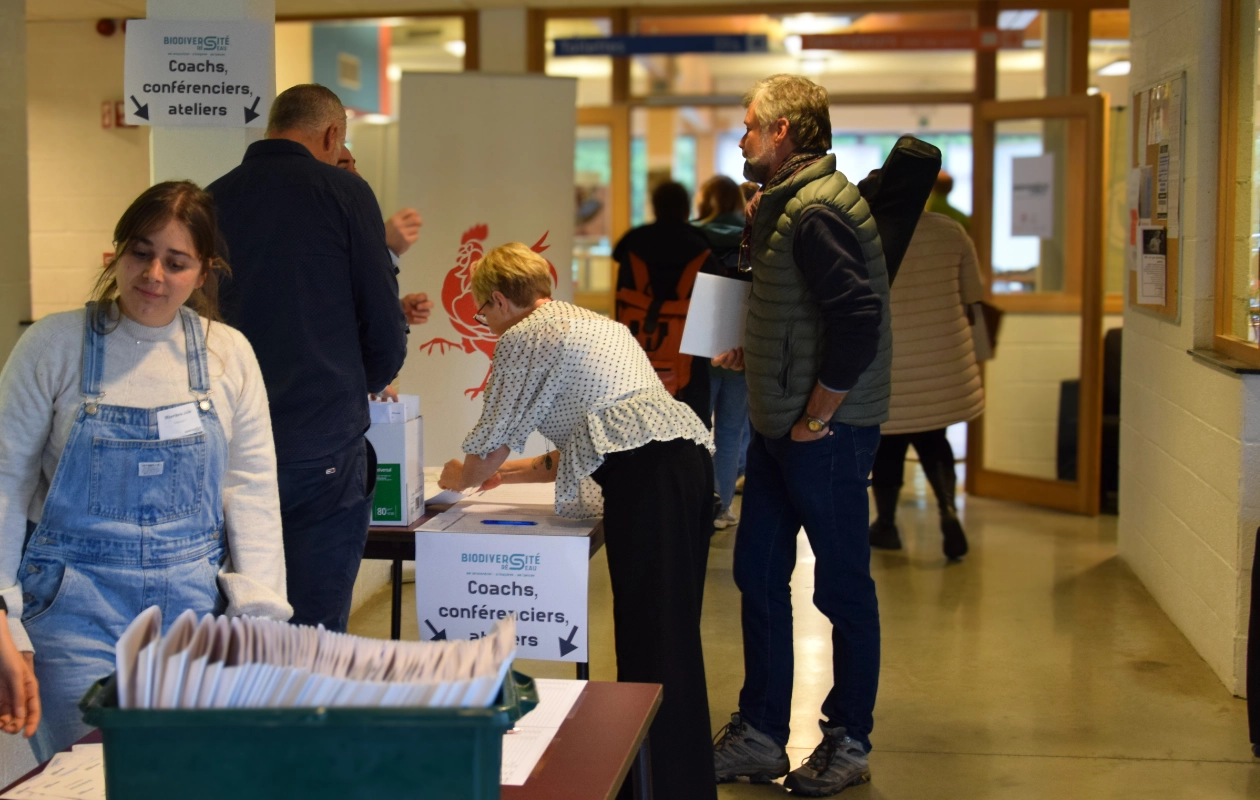 Entrée du bâtiment où se tient la journée des rencontres du réseau BiodiverCité. Les participants récupèrent leur dossier et programme. Plusieurs personnes sont présentes, certaines debout et d'autres interagissant avec des tables où se trouvent les documents. Des panneaux indiquent 'Coachs, conférenciers, ateliers' et 'Réseau BiodiverCité'.