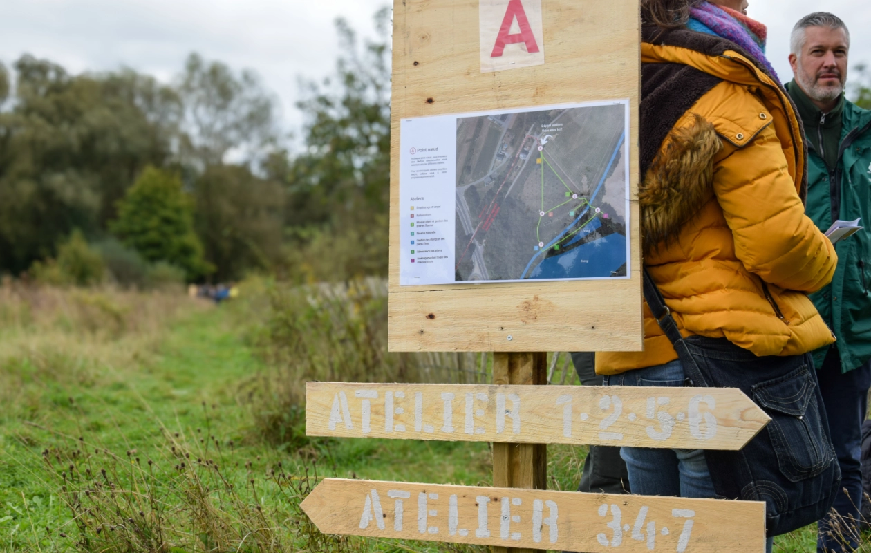 L'image montre des panneaux en bois indiquant la direction des différents ateliers lors des visites de terrain de la journée des Rencontres du Réseau BiodiverCité. Le panneau comporte un 'A' en haut, une carte au milieu, et deux panneaux directionnels en bas : 'ATELIER 1-2-5-6' et 'ATELIER 3-4-7'. Des personnes se tiennent à côté, avec de la verdure en arrière-plan.