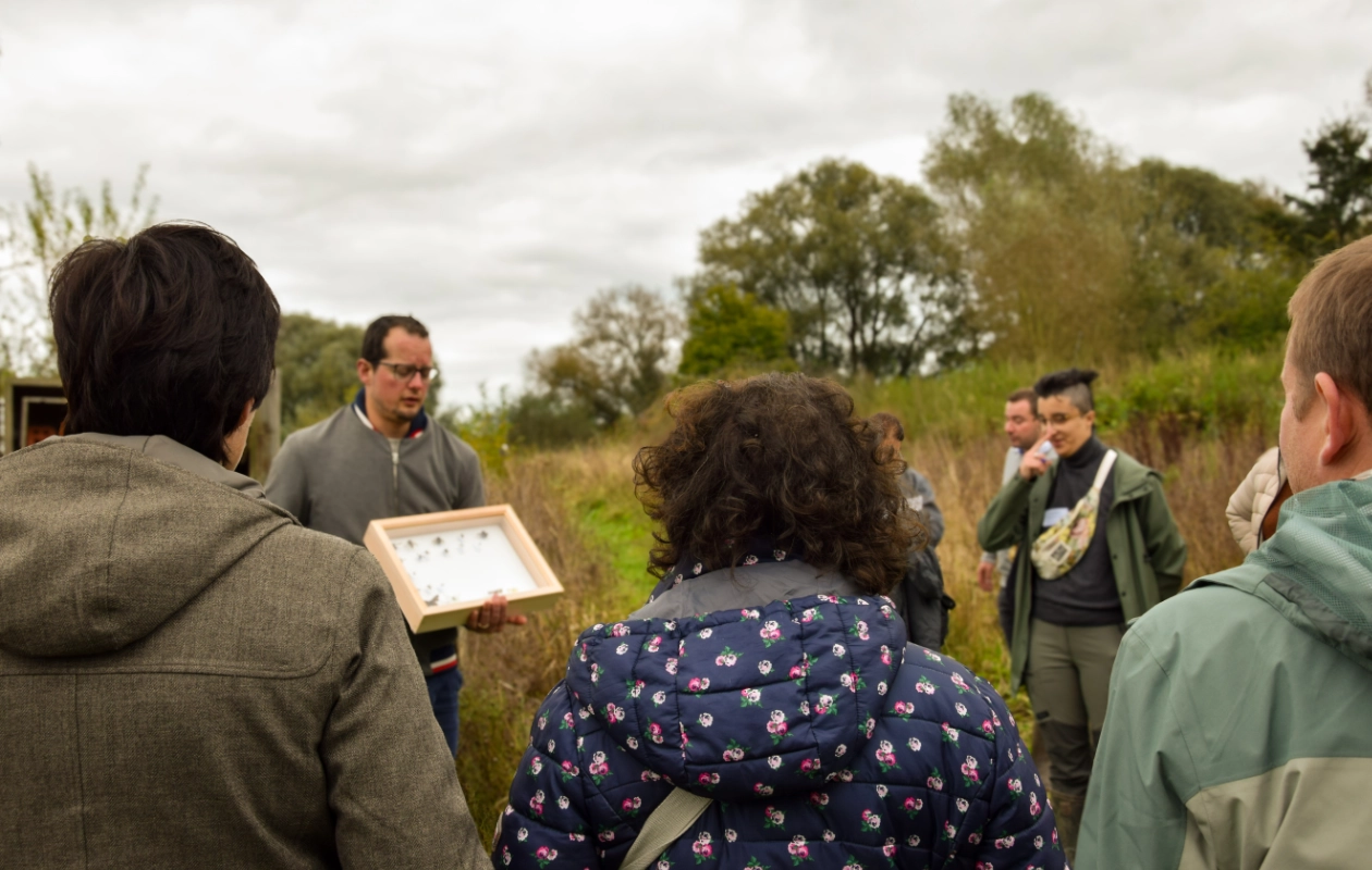 L'image montre un groupe de personnes debout en extérieur dans la réserve de l'Escaille, lors d'un atelier pendant les visites de terrain de la journée des Rencontres du Réseau BiodiverCité. Une personne tient une vitrine avec des spécimens, probablement des insectes, que le groupe observe. En arrière-plan, on voit des arbres et de la végétation sous un ciel couvert.