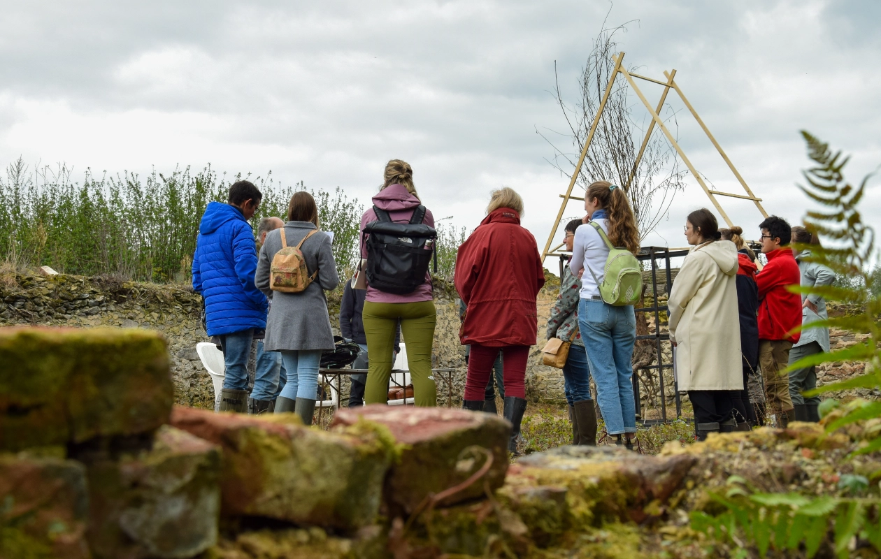 L'image montre un groupe de personnes debout en extérieur dans la réserve de l'Escaille, lors d'un atelier pendant les visites de terrain de la journée des Rencontres du Réseau BiodiverCité. Le groupe, tourné dos à la caméra, semble écouter quelqu'un.