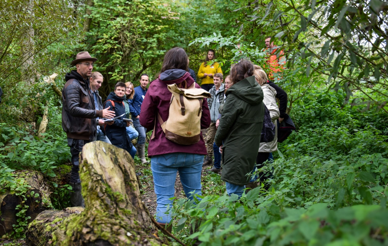 L'image montre un groupe de personnes debout en extérieur dans la réserve de l'Escaille, lors d'un atelier pendant les visites de terrain de la journée des Rencontres du Réseau BiodiverCité. Le groupe, entouré de végétation dense, écoute un expert.