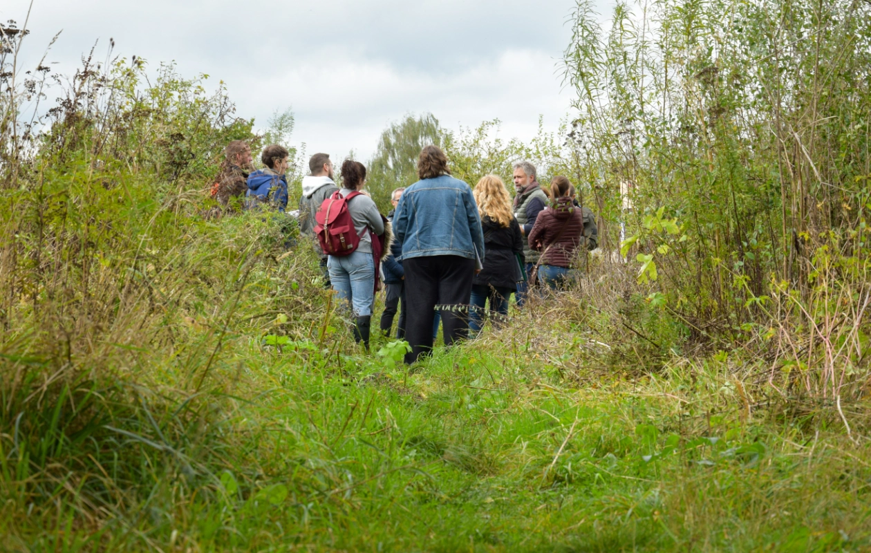 L'image montre un groupe de personnes debout sur un chemin herbeux et envahi par la végétation, entouré de hautes plantes, lors d'un atelier pendant les visites de terrain de la journée des Rencontres du Réseau BiodiverCité. Le groupe semble écouter quelqu'un.