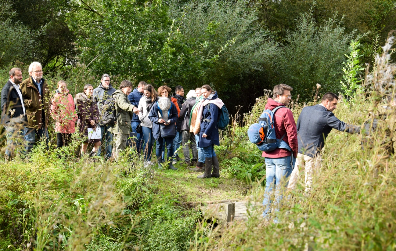 L'image montre un groupe de personnes debout sur un chemin herbeux entouré de végétation dense et d'arbres, lors d'un atelier pendant les visites de terrain de la journée des Rencontres du Réseau BiodiverCité. Le groupe semble écouter quelqu'un, certains observant la végétation, tandis que d'autres discutent ou regardent autour d'eux.