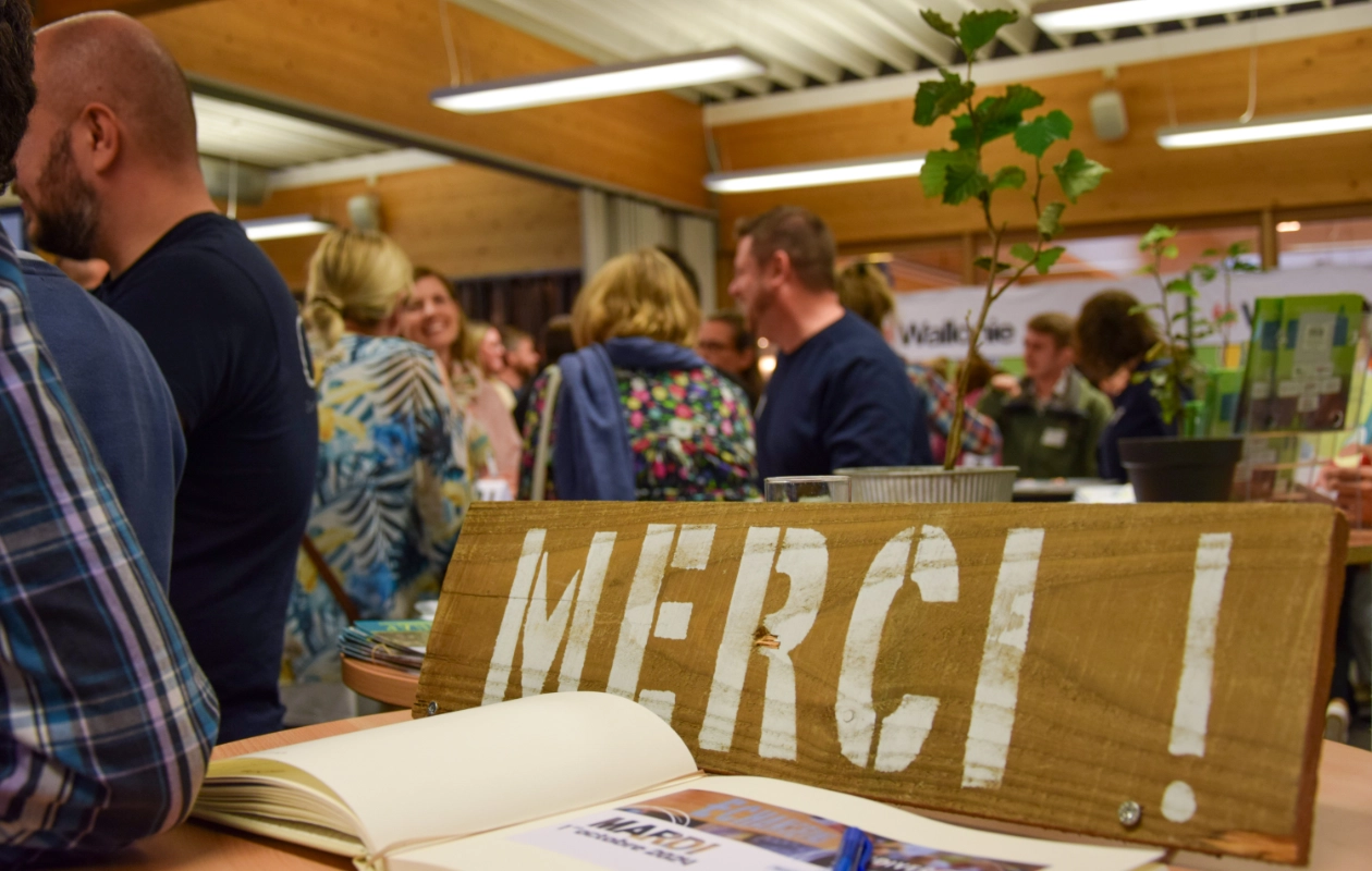 L'image montre une plaque en bois avec l'inscription "MERCI !" placée sur un mange-debout. En dessous de la plaque, un livre d'or est ouvert, révélant du texte et des images. En arrière-plan, plusieurs participants de la journée des Rencontres du Réseau BiodiverCité sont rassemblés et discutent de l'événement qui semble s'achever.