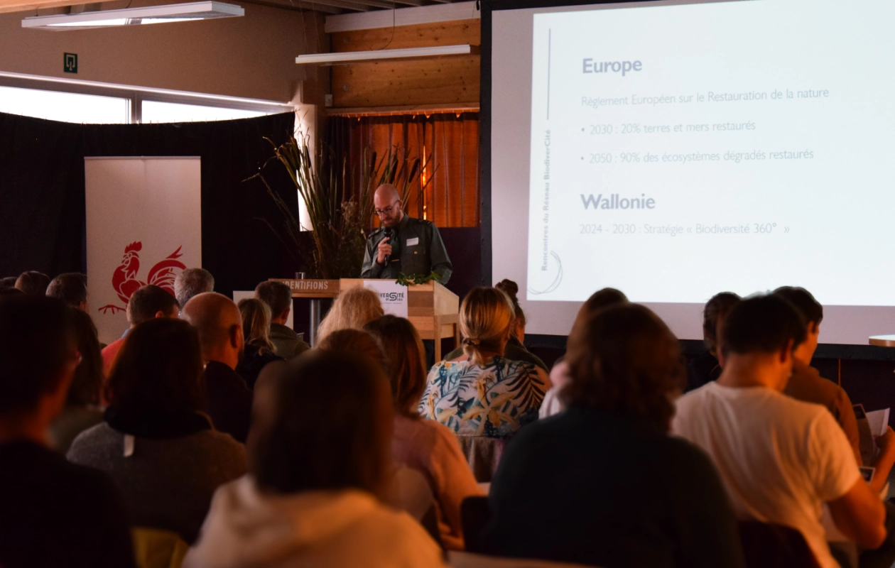 Conférence lors de la journée des rencontres du réseau BiodiverCité donnée par un agent du DNF. Le public est assis et écoute attentivement. La diapositive à l'écran mentionne des règlements et des stratégies pour la restauration de la nature en Europe et en Wallonie.