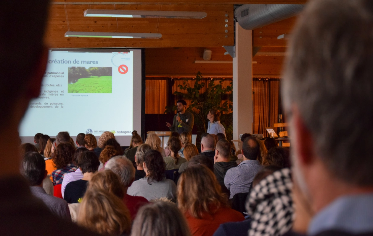Conférence lors de la journée des rencontres du réseau BiodiverCité. Charlotte Busetti du Parc Naturel Viroin-Hermeton et Parc National de l'Entre-Sambre-et-Meuse, ainsi que Luca Fagan de Natagora, présentent sur le thème 'Mares agricoles et forestières, différences et complémentarités pour un réseau fonctionnel'. Le public est assis et écoute attentivement.