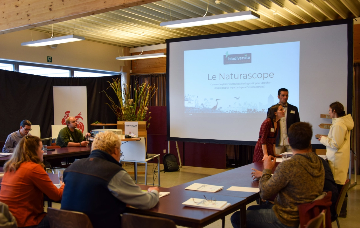 L'image montre un atelier sur le Naturascope, un outil d'autodiagnostic de la biodiversité pour les communes, mené par deux agents de la FRW pendant la journée des Rencontres du Réseau BiodiverCité. À l'avant de la salle, un grand écran affiche la présentation. Les deux protagonistes sont debout près de l'écran et interagissent avec les participants, assis face à eux autour de tables.