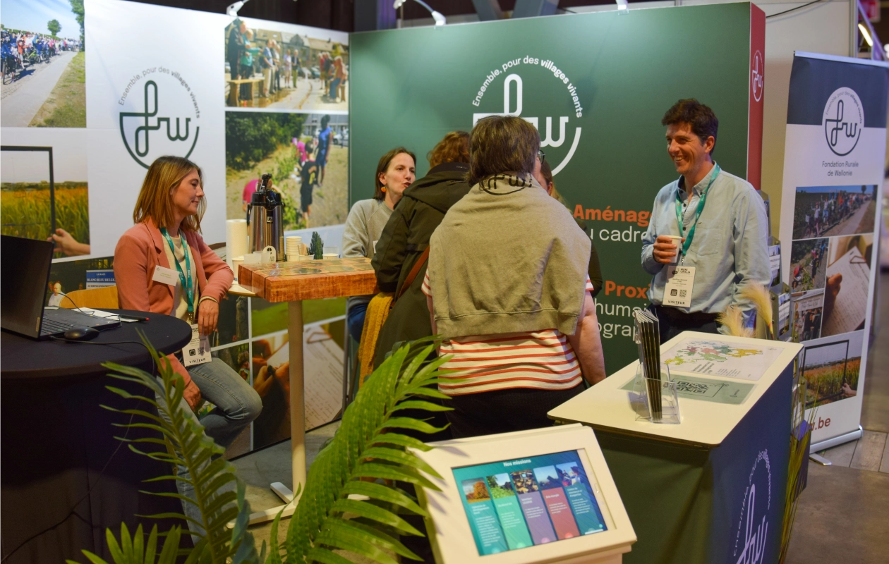 Stand de la Fondation Rurale de Wallonie (FRW) au salon Municipalia 2025. Plusieurs personnes discutent autour d'une table haute. Le stand est décoré avec des affiches etpanneaux présentant les activités de la FRW.