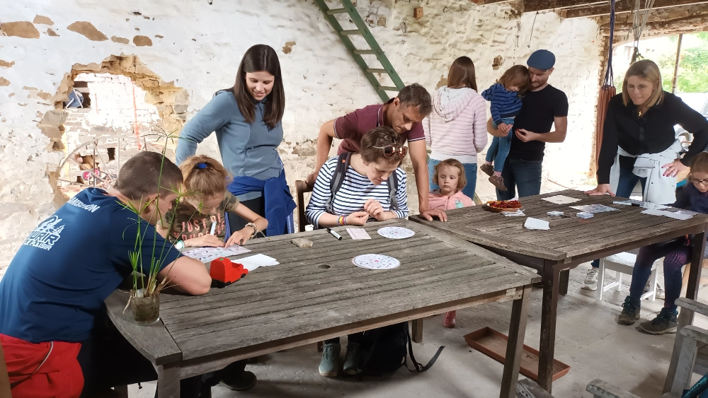Groupe de personnes rassemblées autour de deux tables en bois dans un cadre intérieur rustique. Les murs sont en pierre et une échelle est appuyée contre l'un des murs. Certaines personnes sont assises tandis que d'autres sont debout, et elles semblent être engagées dans diverses activités impliquant du papier et d'autres matériaux sur les tables.