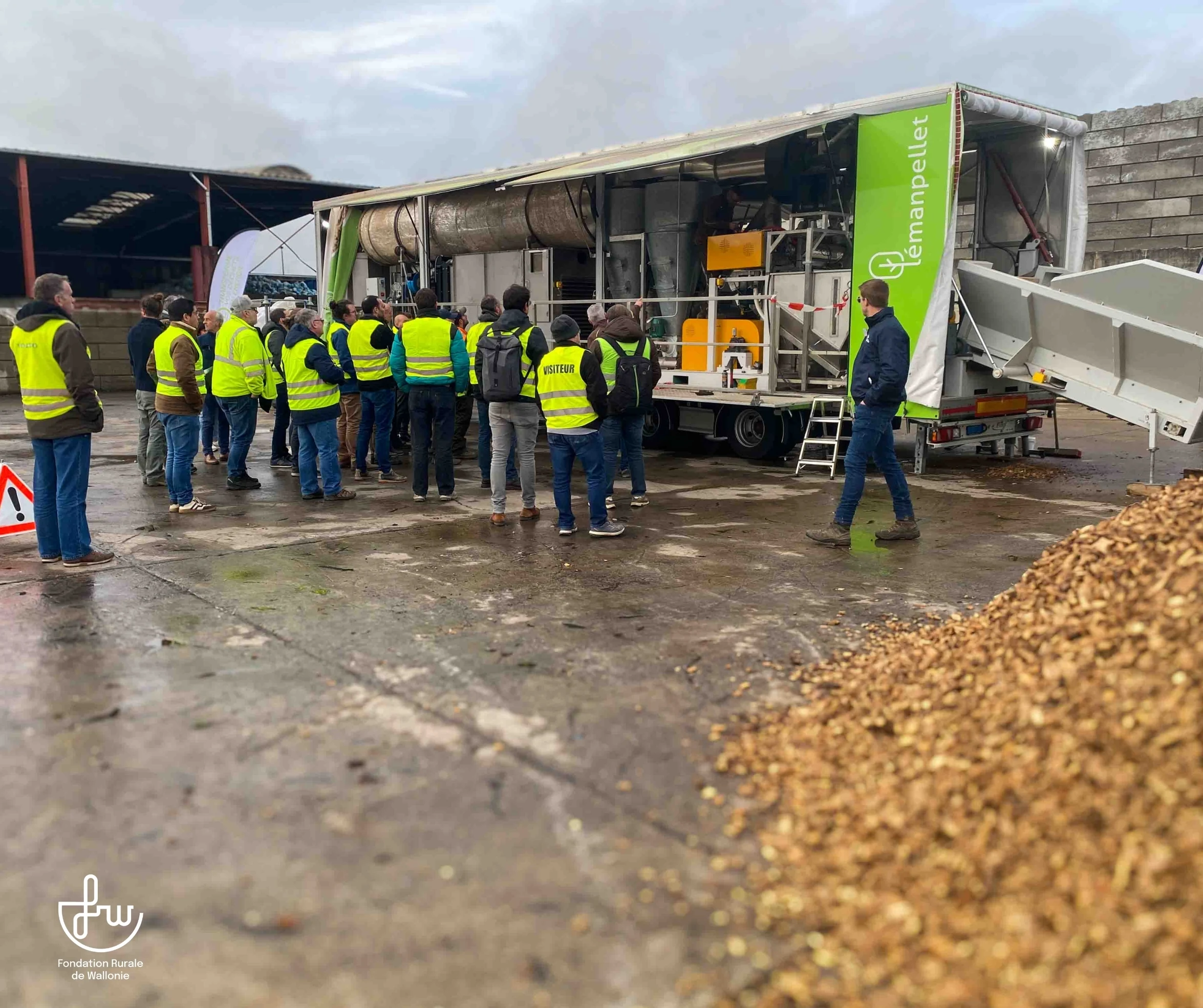 Groupe de personnes portant des gilets jaunes rassemblées autour d'une machine mobile. Celle-ci semble être liée au traitement de la biomasse, comme l'indique l'inscription 'Lémanpellet' sur le côté de la remorque et le tas de copeaux de bois visible au premier plan. Le cadre semble être un site industriel ou agricole, comme le suggèrent les hangars visibles en arrière-plan.