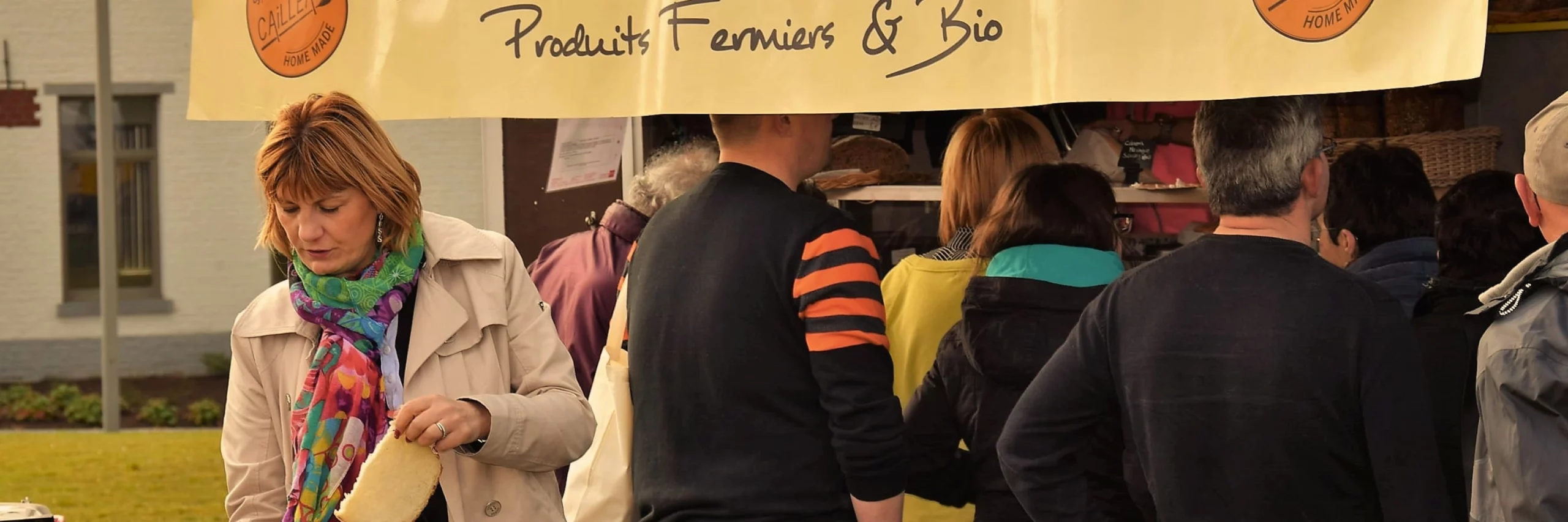 Des personnes qui font la file devant une remorque aménagée en échoppe pour vendre des produits fermiers et bio. Au premier plan, une maman qui donne une tranche de pain fraîchement acheté à son enfant.