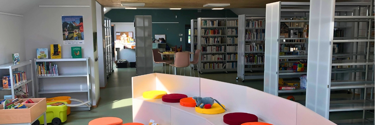 Vue intérieure d'une bibliothèque et d'un espace de lecture. Des étagères remplies de livres, une zone de sièges avec des coussins ronds colorés, et une petite table avec des jouets.