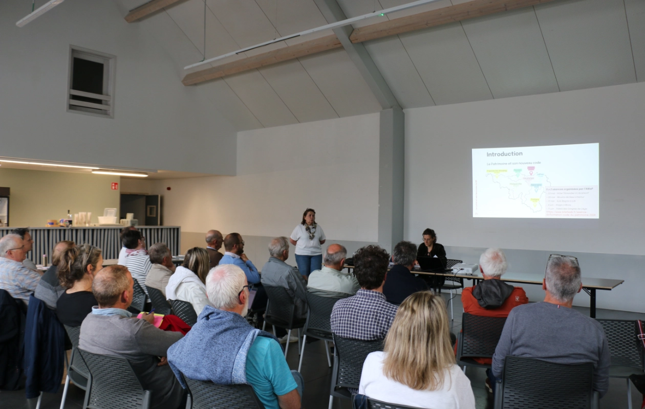 Formation sur le nouveau code wallon du patrimoine en intérieur avec une présentation. Deux personnes sont à l’avant, l’une debout parlant et l’autre assise avec un ordinateur portable. Environ vingt personnes sont assises face aux présentateurs. Un écran de projection affiche une diapositive intitulée ‘Introduction’, accompagnée d'une carte de la Wallonie.