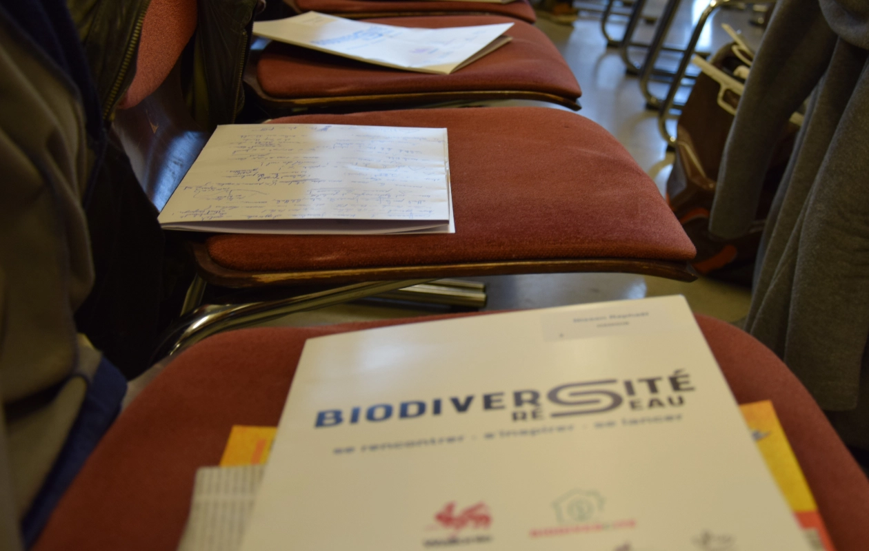 Rangée de chaises avec des documents posés dessus. Le document en focus porte le titre 'BIODIVERSITÉ RÉSEAU' et inclut des logos et du texte en dessous. Des notes manuscrites sont visibles sur un autre document en arrière-plan.