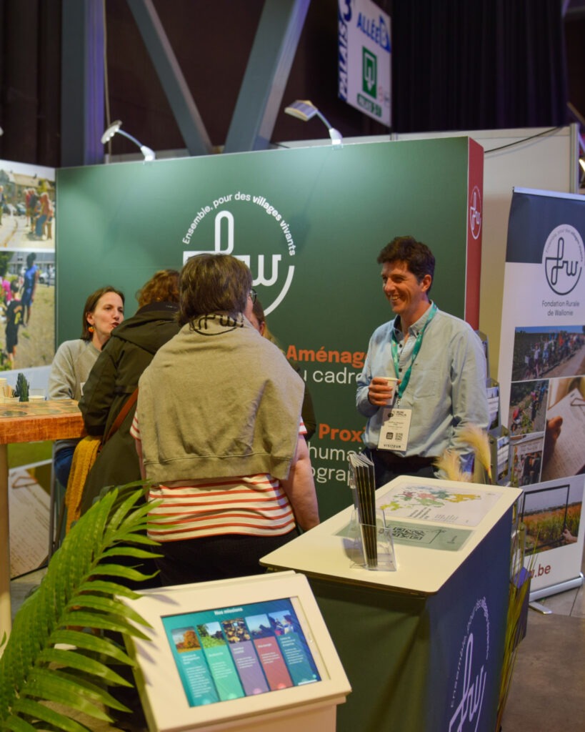 Stand de la Fondation Rurale de Wallonie (FRW) au salon Municipalia 2025. Plusieurs personnes discutent autour d'une table haute. Le stand est décoré avec des affiches et panneaux présentant les activités de la FRW.