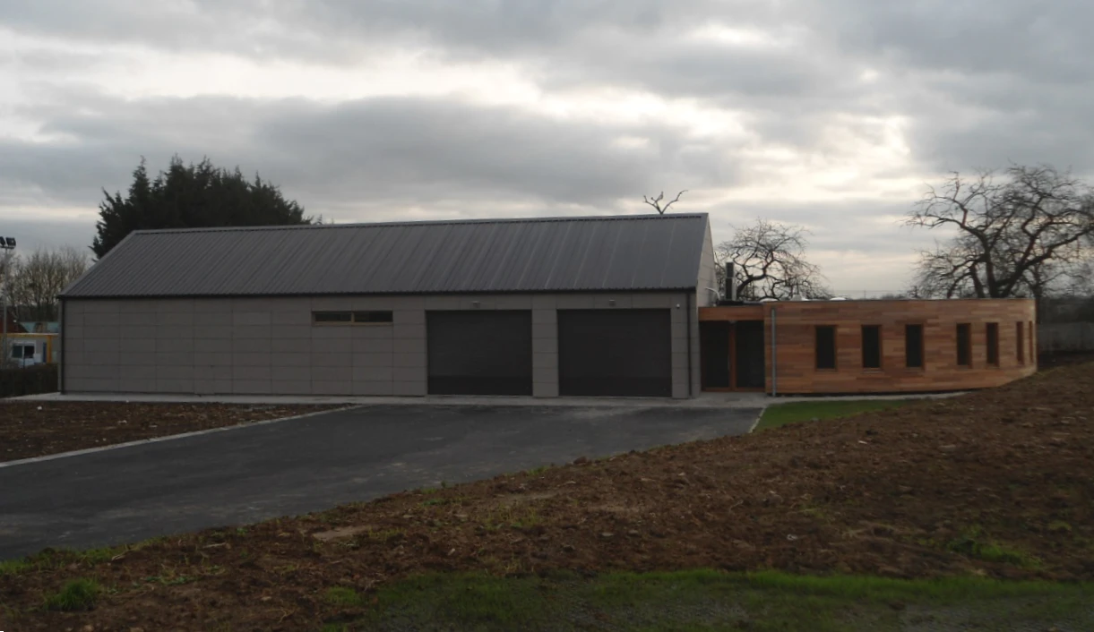 Bâtiment moderne avec finitions extérieures en métal et bois, doté d'un grand toit en métal gris foncé et de trois grandes portes de garage. La section gauche est en panneaux métalliques, tandis que la section droite présente un bardage en bois. Le paysage environnant est récemment aménagé avec des zones de gazon et de sol nu, des arbres en arrière-plan, et un ciel nuageux.