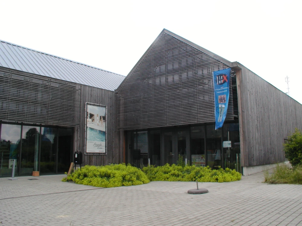 Bâtiment moderne avec un bardage en bois et un toit incliné. L'entrée principale est en verre, et la zone devant le bâtiment est pavée de briques. Une bannière bleue est accrochée sur le côté droit du bâtiment, et une affiche est visible près de l'entrée à gauche. L'environnement autour du bâtiment comprend des buissons et de l'herbe.