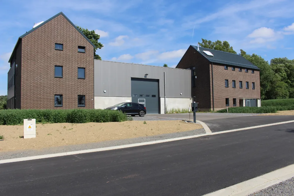 Atelier rural à Jalhay composé de deux structures en briques reliées par une section centrale avec de grandes portes de garage. Une voiture noire est garée devant la section centrale. L'environnement autour des bâtiments est aménagé avec de la verdure et des routes pavées. Le ciel est dégagé avec quelques nuages visibles.