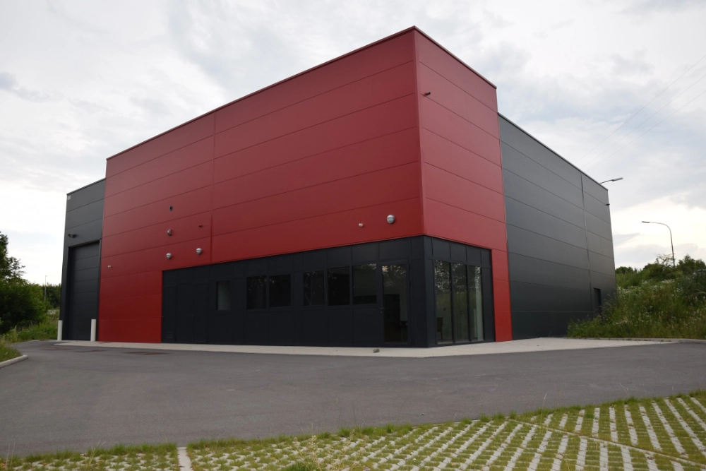 Bâtiment industriel moderne avec une façade rouge et noire. Le bâtiment a un toit plat et plusieurs fenêtres sur le devant. La zone pavée devant le bâtiment semble faire partie du parking ou de l'allée. Les environs comprennent de la verdure et des arbres, indiquant qu'il est situé dans une zone semi-rurale ou suburbaine.