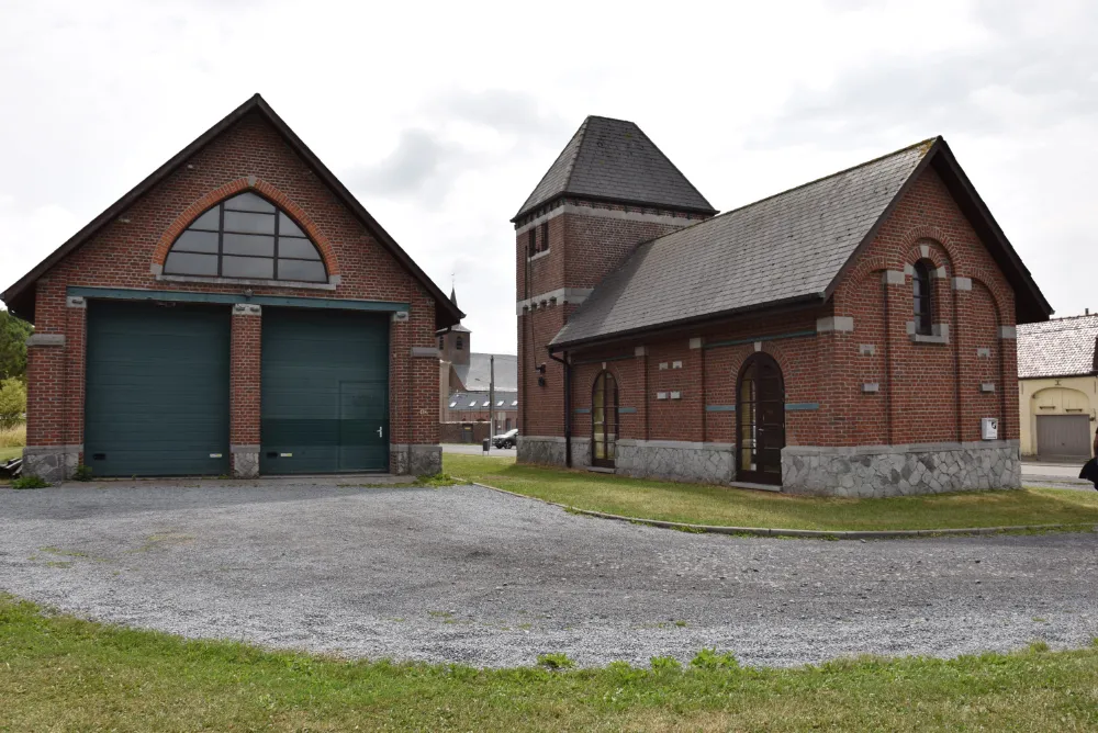 Atelier rural composé de deux bâtiments en briques avec des toits en pente. Le bâtiment de gauche a deux grandes portes de garage vertes et une fenêtre en arc au-dessus. Le bâtiment de droite a une porte et des fenêtres en arc plus petites, avec une structure en forme de tour au milieu. Les bâtiments sont entourés d'une allée en gravier et de zones herbeuses.