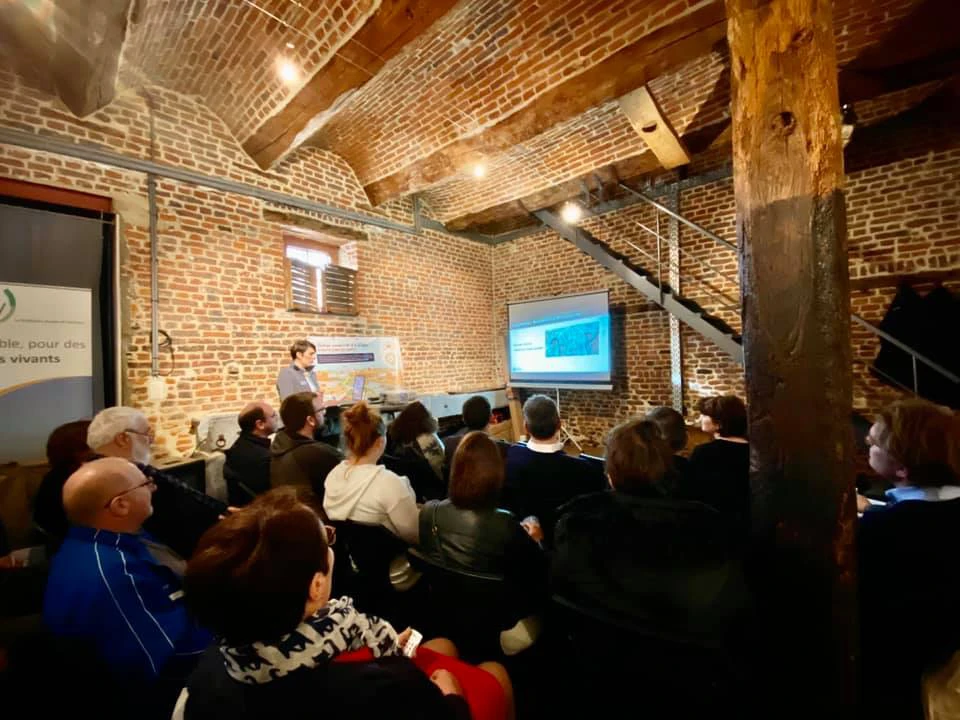 Groupe de personnes assises dans une salle avec des murs en briques et des poutres en bois, assistant à une présentation. Le présentateur se tient à l'avant près d'un écran de projection affichant une image ou une diapositive. Il y a également une bannière sur le côté gauche de la salle.