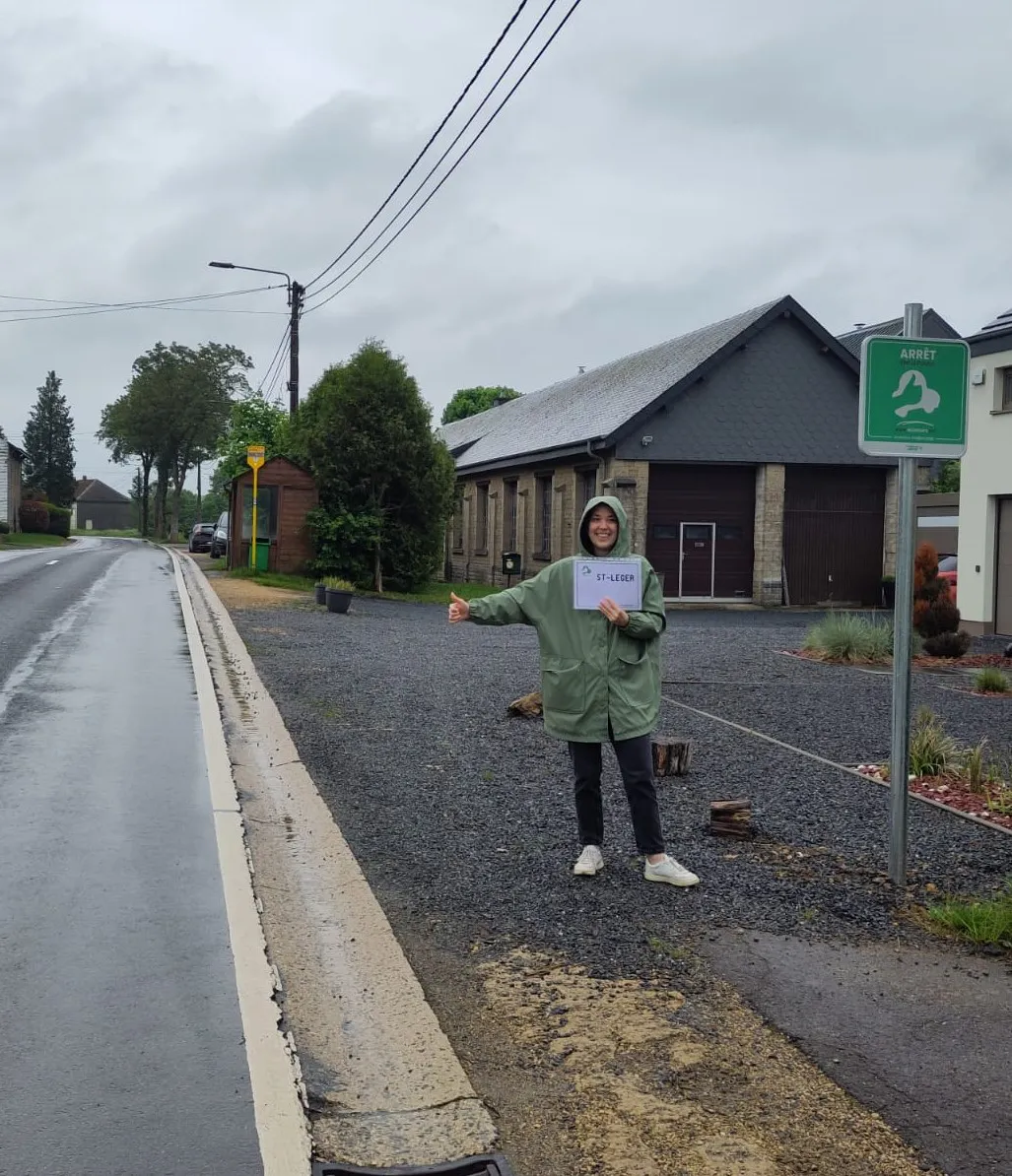 Personne faisant de l'auto-stop : Une personne se tenant sur le bord de la route, levant le pouce, ce qui est couramment associé à l'auto-stop. La personne porte une veste verte et un pantalon noir. Un panneau à proximité indique "ARRÊT" avec une icône de main levant le pouce, suggérant qu'il s'agit probablement d'un endroit désigné pour l'auto-stop.