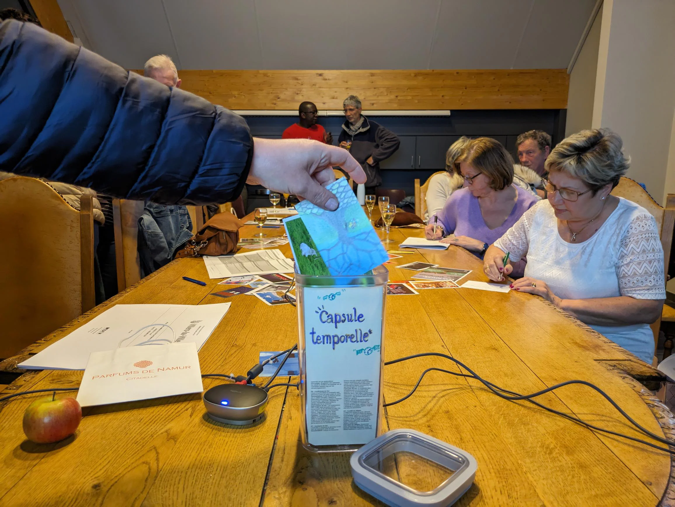 L'image montre un groupe de personnes assises autour d'une table en bois, occupés à discuter, rédiger ou dessiner. En amorce, une main place un objet, vraisemblablement un message papier, dans une boîte étiquetée "Capsule temporelle".