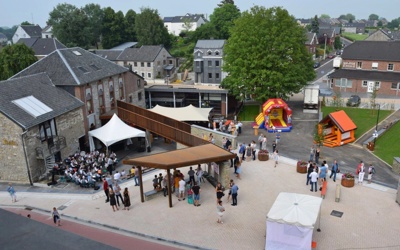 Un événement en plein air avec plusieurs groupes de personnes rassemblées dans différentes zones. Il y a des tentes, une scène avec un groupe ou un orchestre en train de jouer, et divers stands ou kiosques. En arrière-plan, il y a deux châteaux gonflables et quelques bâtiments. Le cadre semble être une cour ou un espace ouvert entouré de bâtiments et d'arbres.
