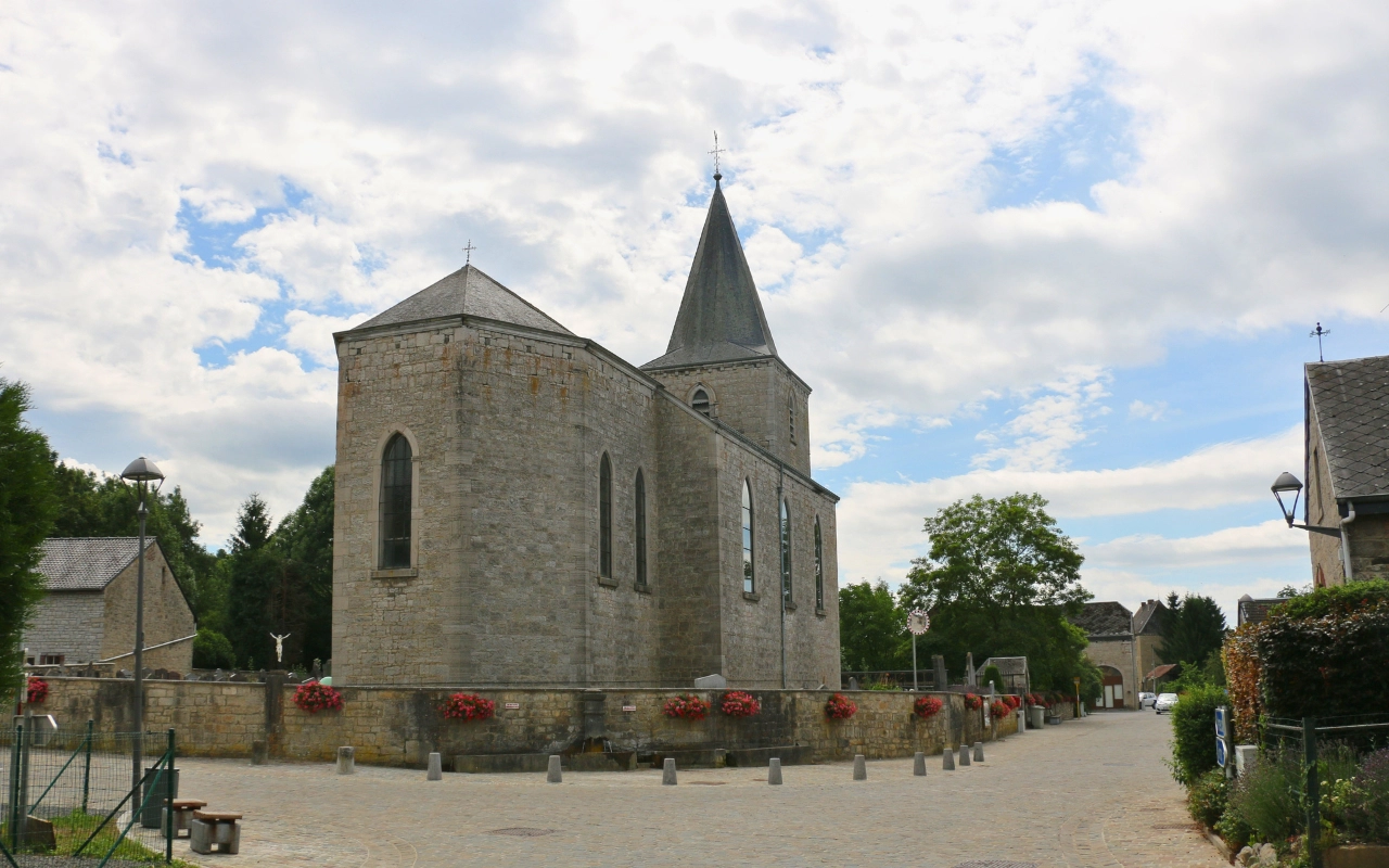 Une église en pierre avec un clocher pointu dans un petit village. L'église est entourée d'une rue pavée de pierres et comporte des jardinières avec des fleurs rouges le long de son périmètre. Il y a d'autres bâtiments et des arbres visibles en arrière-plan sous un ciel partiellement nuageux.