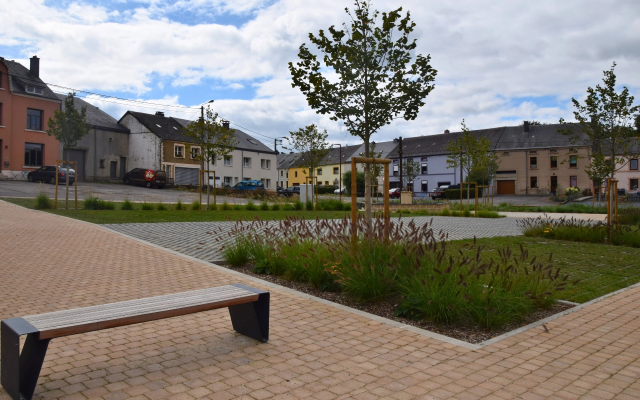 Une place publique avec une zone pavée, plusieurs jeunes arbres plantés dans des parcelles désignées, et un banc au premier plan. En arrière-plan, il y a des bâtiments résidentiels de différentes couleurs et designs. Le ciel est partiellement nuageux.