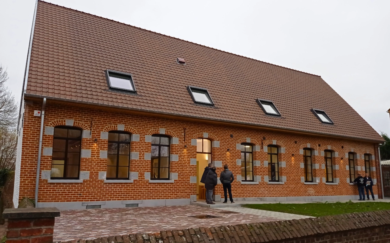 L'image montre un bâtiment en briques avec un toit incliné et plusieurs fenêtres. Des personnes se tiennent près de l'entrée du bâtiment. Le bâtiment a une zone pavée à l'avant et de l'herbe sur le côté.