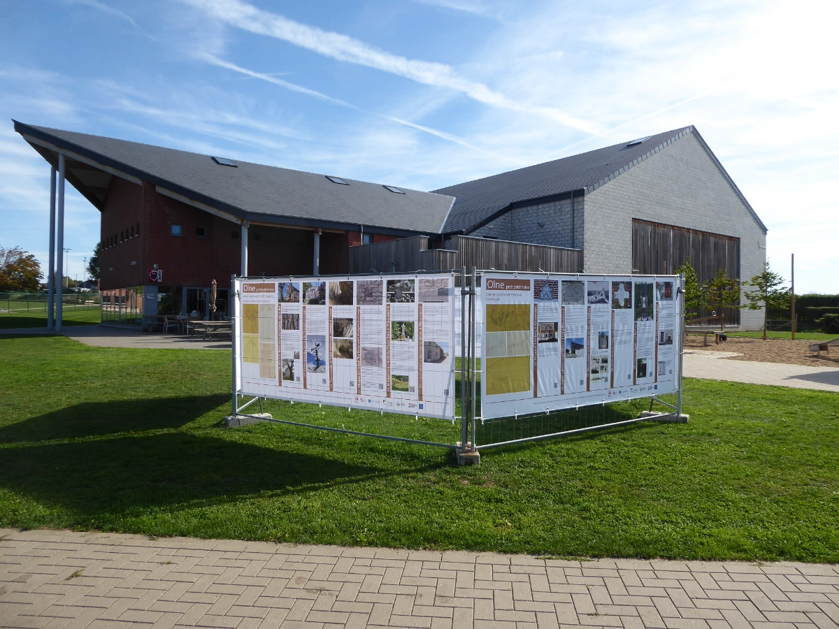 Un affichage extérieur de panneaux d'information installés sur une zone herbeuse. L'affichage est encadré par une structure métallique et se compose de plusieurs panneaux avec du texte et des images. En arrière-plan, il y a deux bâtiments modernes avec des toits inclinés, l'un en brique et l'autre en matériau gris. Le ciel est dégagé avec quelques nuages effilochés, et il y a un chemin pavé au premier plan.