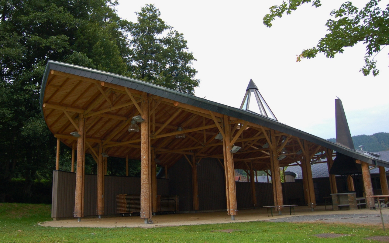 L'image montre une structure de pavillon en bois avec un toit courbé soutenu par plusieurs colonnes en bois. Le pavillon est ouvert sur les côtés et possède une zone partiellement fermée à l'arrière. Il y a des arbres en arrière-plan, et le sol autour du pavillon est pavé.