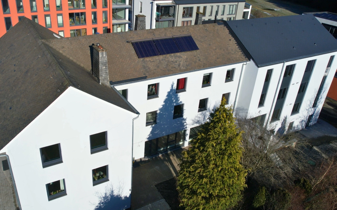 L'image montre un bâtiment à plusieurs étages avec un toit incliné et plusieurs fenêtres. Il y a une série de panneaux solaires sur le toit du bâtiment. Le bâtiment est entouré d'arbres et d'autres bâtiments en arrière-plan.