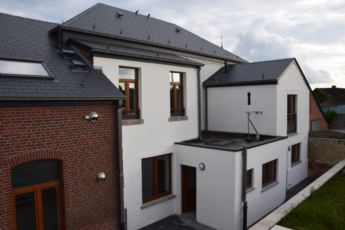 L'image montre une maison avec une combinaison de murs extérieurs en briques peintes en blanc et en briques rouges. Le toit est en bardeaux gris foncé, et il y a plusieurs fenêtres avec des cadres en bois. Il y a aussi un petit balcon sur le côté droit de la maison. Le ciel en arrière-plan est partiellement nuageux.
