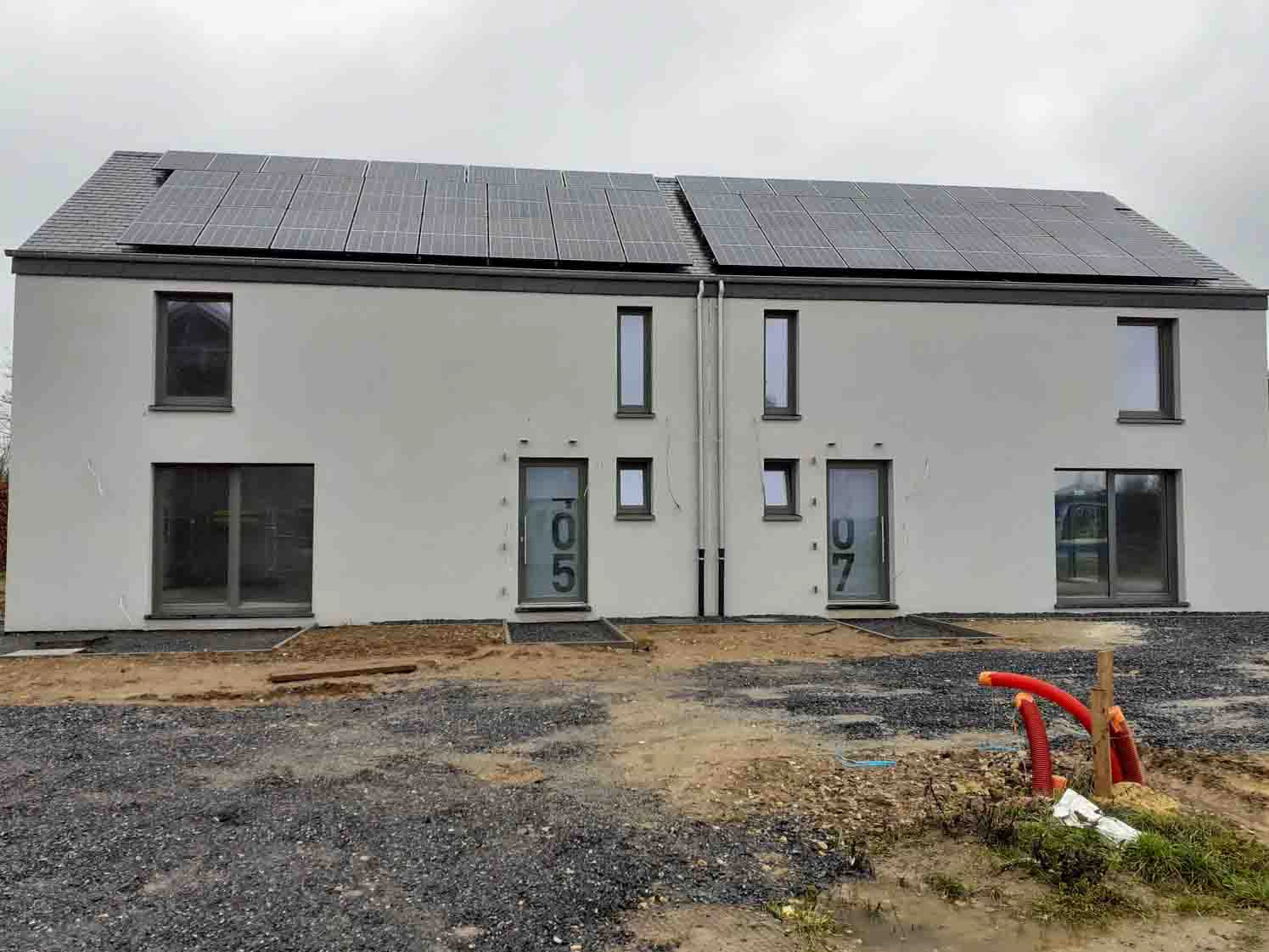 L'image montre un duplex moderne à deux étages avec des panneaux solaires installés sur le toit. Le bâtiment a une façade blanche et de grandes fenêtres. Chaque unité a sa propre entrée, marquée avec les numéros "05" et "07". La zone devant le bâtiment semble être en cours de construction ou d'aménagement paysager, avec du gravier et du sol exposé. Il y a aussi quelques tuyaux rouges visibles au premier plan.