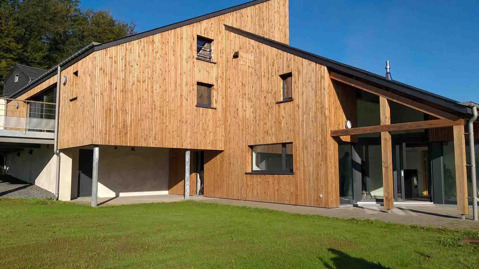 L'image montre une maison moderne en bois avec un design angulaire et multi-niveaux. L'extérieur est revêtu de planches de bois verticales, et la maison dispose de grandes fenêtres et d'un toit incliné. Il y a une pelouse verte devant la maison, et une partie de la structure est surélevée sur des pilotis. Le ciel est dégagé et bleu, indiquant une journée ensoleillée.