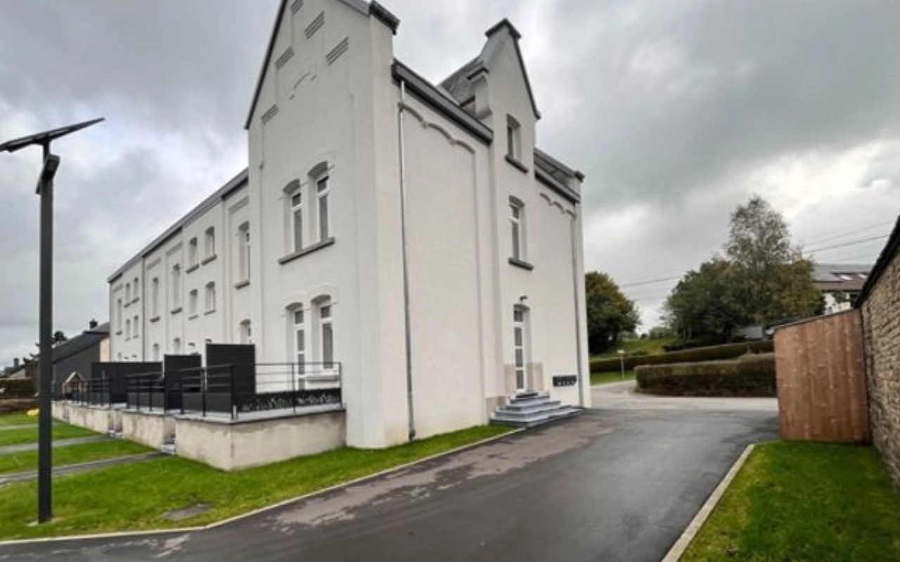 L'image montre un grand bâtiment blanc à deux étages avec un toit fortement incliné et plusieurs fenêtres. Le bâtiment semble être résidentiel ou peut-être une structure institutionnelle. Il y a une allée pavée menant à l'entrée du bâtiment, qui a des marches et un petit palier. La zone environnante comprend des pelouses bien entretenues et quelques arbres en arrière-plan. Il y a aussi un abri en bois sur le côté droit de l'image et un lampadaire moderne sur le côté gauche. Le ciel est couvert avec des nuages gris.