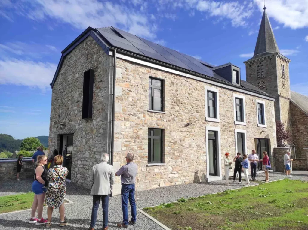 L'image montre un groupe de personnes debout à l'extérieur d'un bâtiment en pierre avec des panneaux solaires sur le toit. Le bâtiment a plusieurs fenêtres et portes, et il y a une tour d'église attachée. Le ciel est dégagé avec quelques nuages, et la zone environnante semble être herbeuse avec quelques chemins en gravier.
