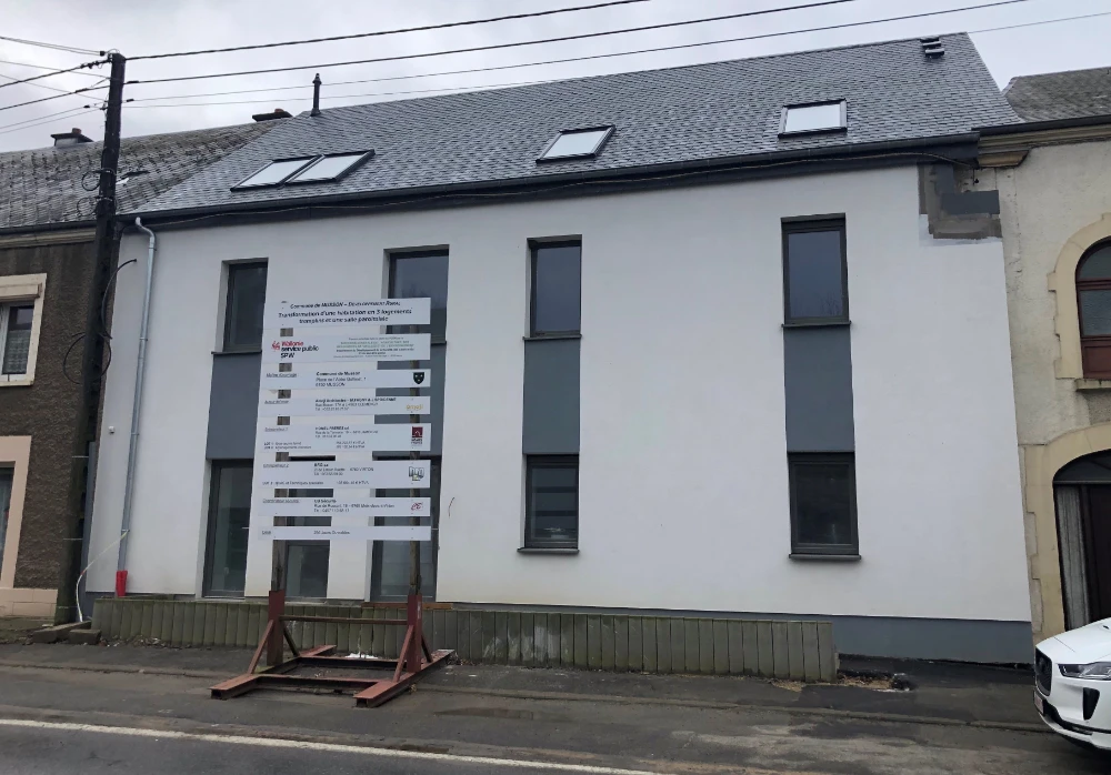 L'image montre un bâtiment avec un toit gris et des murs blancs. Il y a trois fenêtres verticales sur la façade du bâtiment, et un panneau d'affichage devant. Le panneau contient plusieurs lignes de texte, probablement liées à des informations de construction ou de rénovation.