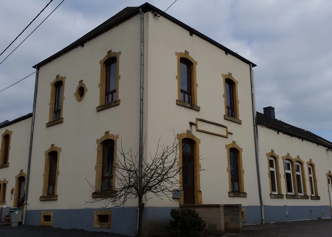 L'image montre un bâtiment à deux étages avec des murs de couleur crème et des bordures jaunes autour des fenêtres et des portes. Le bâtiment a plusieurs fenêtres hautes et étroites, dont certaines sont en arc en haut. Il y a un petit arbre sans feuilles devant le bâtiment, et le ciel semble être nuageux.
