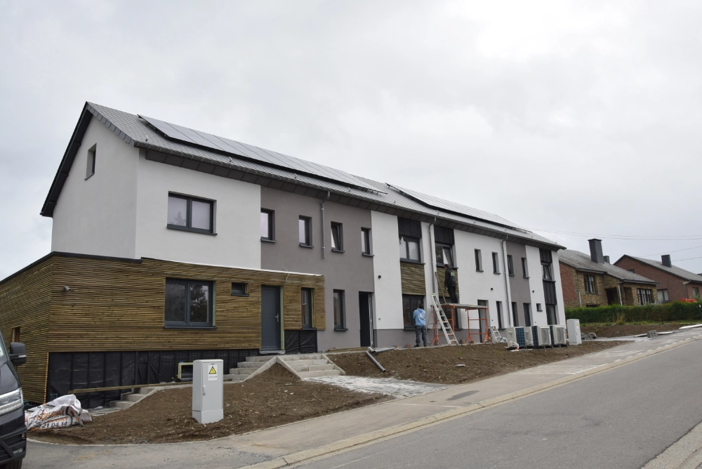 L'image montre une rangée de maisons de ville modernes en cours de construction ou de rénovation. Les bâtiments ont des extérieurs mêlant blanc et gris avec des accents de panneaux en bois. Des panneaux solaires sont installés sur les toits. On peut voir des équipements et des matériaux de construction, y compris des échelles et des échafaudages, indiquant des travaux en cours. La zone devant les maisons semble être préparée pour l'aménagement paysager ou d'autres travaux de construction.
