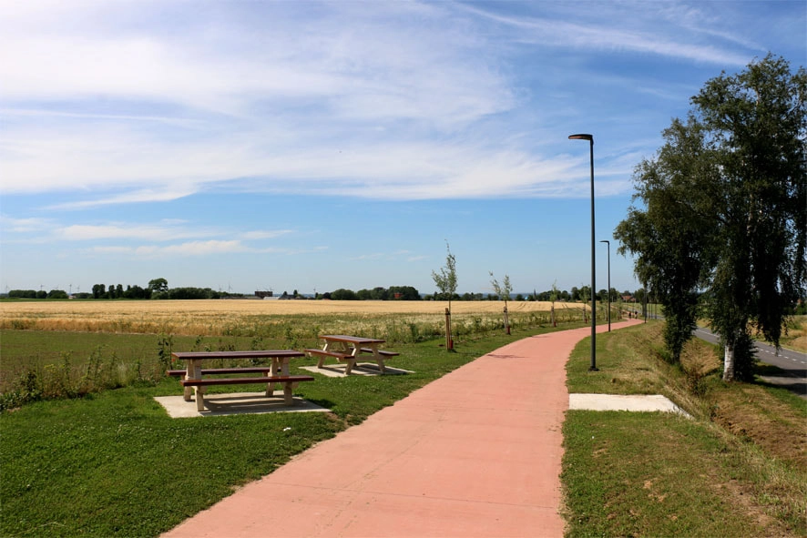 Chemin pavé avec tables de pique-nique : Un chemin pavé traversant une zone herbeuse avec des tables de pique-nique sur le côté gauche. Il y a des lampadaires le long du chemin, et des champs s'étendent au loin sous un ciel partiellement nuageux. Des arbres et des arbustes sont visibles de chaque côté du chemin.