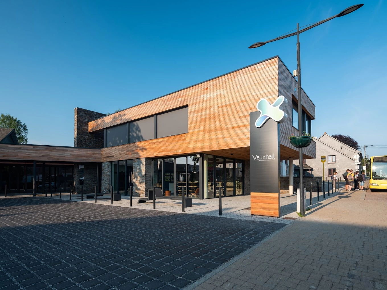 L'image montre un bâtiment moderne avec une façade en bois et de grandes fenêtres vitrées. Il y a un panneau devant le bâtiment avec le texte "Vauxhall" et un logo au-dessus. Le bâtiment est situé sur une zone pavée avec des bornes devant, et un lampadaire est visible. Un bus jaune peut être vu sur le côté droit de l'image.
