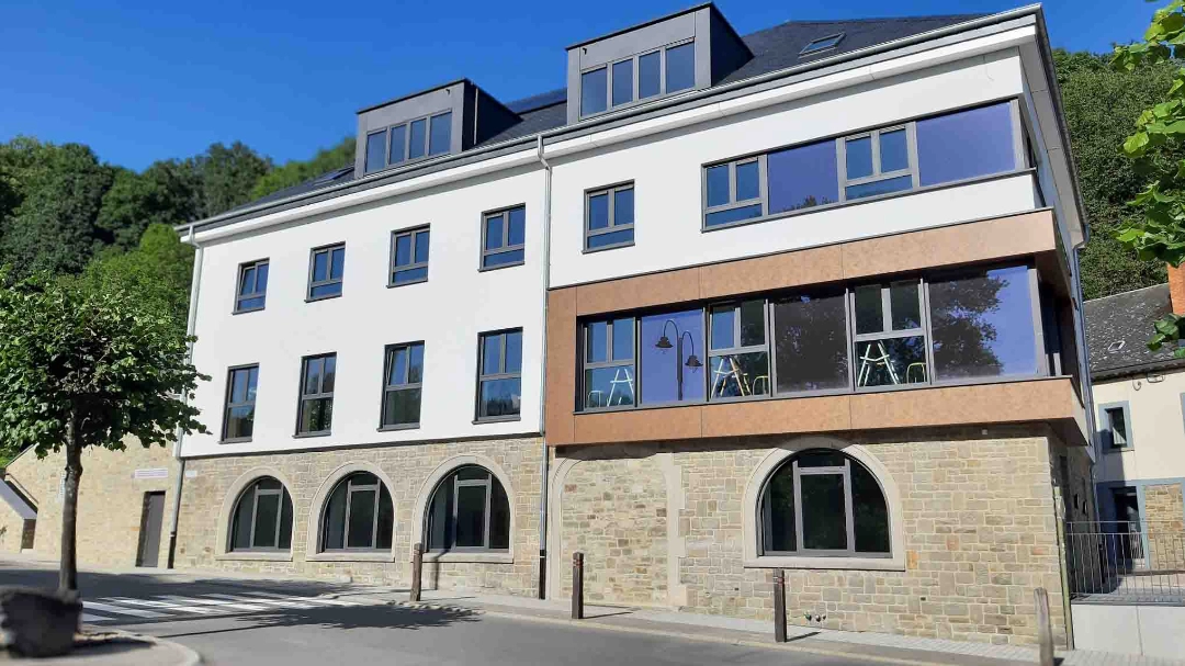L'image montre un bâtiment moderne à plusieurs étages avec une combinaison de murs extérieurs blancs et en pierre. Le bâtiment a de grandes fenêtres à chaque étage, y compris des fenêtres en arc au rez-de-chaussée. Il y a des arbres et une rue devant le bâtiment.