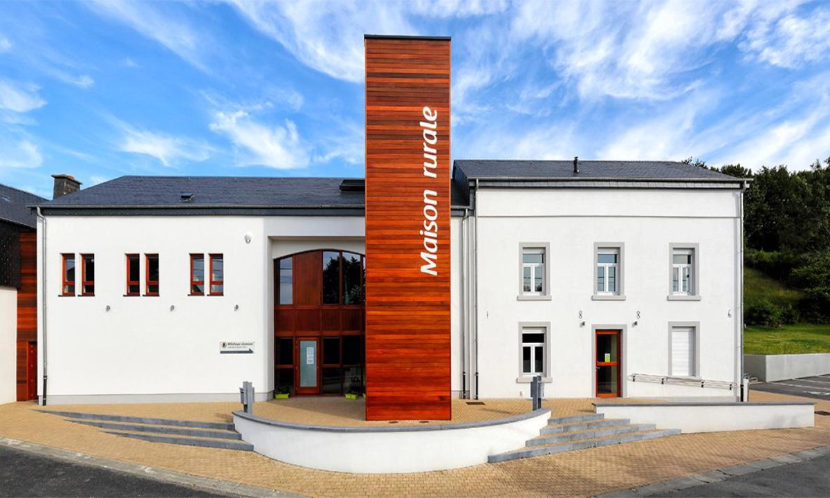 Bâtiment rénové à l'apparence moderne avec une façade en crépi blanc et quelques éléments en bois, notamment la partie centrale avec un bardage surpassant la hauteur du bâtiment avec l'inscription "maison rurale" en verticale.