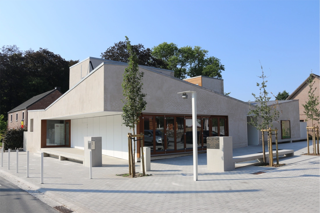 Bâtiment moderne avec un design architectural minimaliste. Le bâtiment a un toit plat et est construit avec des matériaux de couleur claire, une grande baie vitrée sur la façade avant, et l'entrée sur le côté gauche du bâtiment. La zone environnante est pavée, avec des arbres fraîchement plantés.