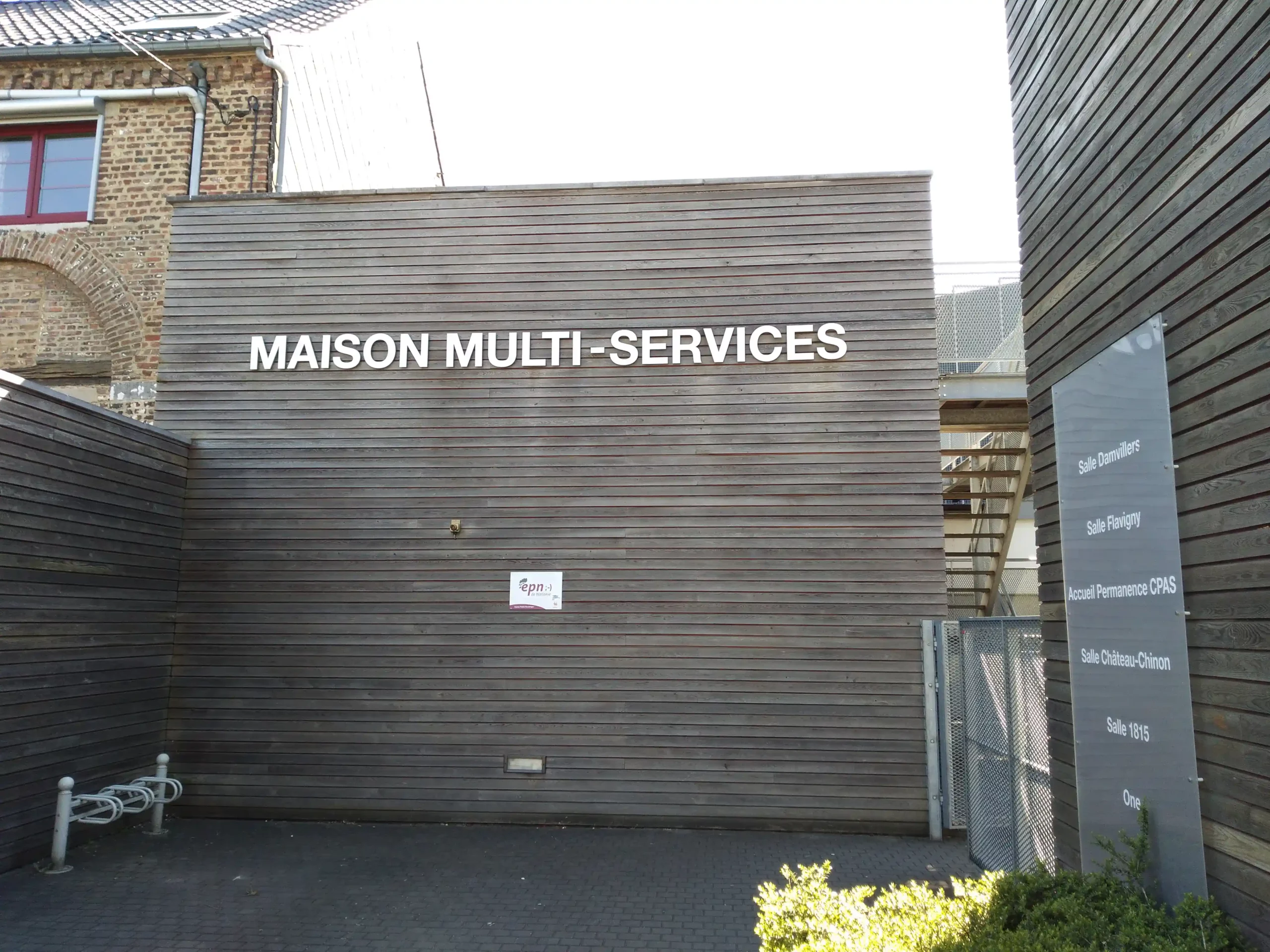 Façade en bardage bois d'un bâtiment avec l'inscription géante "MAISON MULTI-SERVICES". D'autres mentions figurent sur la façade à divers endroits : "Espace public numérique de Wallonie", "Salle Damvillers", "Salle Flavigny", "Accueil Permanences CPAS", "Salle Château-Chinon", "Salle 1815", "ONE".