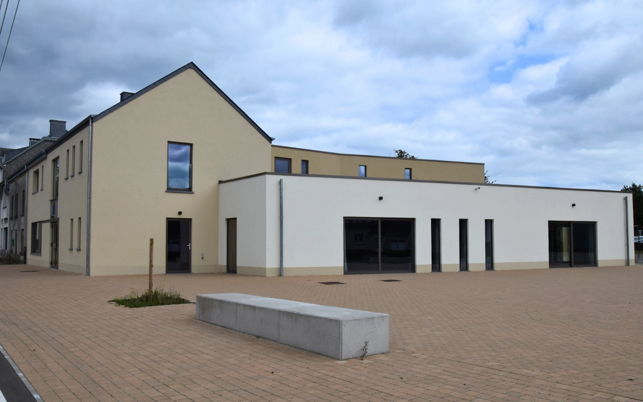 L'image montre un bâtiment moderne avec une façade beige et blanche. Le bâtiment a plusieurs sections, y compris une partie à deux étages sur la gauche et une partie à un étage sur la droite. Il y a plusieurs fenêtres et portes visibles, ainsi qu'une zone pavée ouverte devant le bâtiment avec un banc en béton. Le ciel est nuageux.