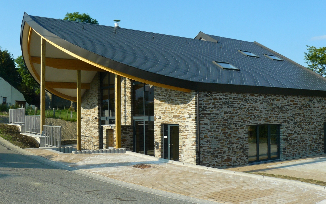 L'image montre un bâtiment moderne avec un design unique de toit courbé. Les murs extérieurs sont en pierre, et il y a de grandes fenêtres et portes vitrées à l'avant. Le toit est de couleur sombre avec plusieurs puits de lumière. Il y a une zone pavée devant le bâtiment, qui semble être une entrée ou une allée. Le bâtiment est situé sur une légère pente, avec des marches et des rampes menant à l'entrée sur un côté.